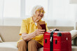 © SHOTPRIME STUDIO - Happy senior woman with passport and travel ticket packed a red suitcase, vacation and health care. Smiling old woman joyfully sitting on the sofa before the trip raised her hands up in joy.