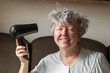 © Kseniia - Smiling senior woman dries her grey hair with a hair dryer. Happy grandma using hair dryer. Active seniors concept.
