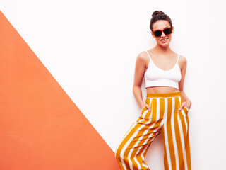 Fashion young beautiful smiling female in trendy summer white top t-shirt and yellow striped pants. Sexy carefree woman posing near wall in studio interior. Positive model. In sunglasses