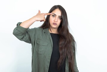 Wall Mural - Unhappy young brunette woman wearing casual clothes over white studio wall makes suicide gesture and imitates gun with hand, curves lips keeps two fingers on temple, shoots, being tired of everything,