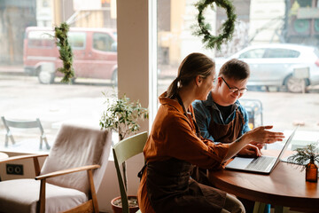 Young woman using laptop while training man with down syndrome to work in cafe