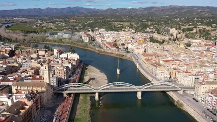 Wall Mural - Aerial view of Catalan city of Tortosa on sunny spring day overlooking reconstructed Bridge of State across Ebro river and Monument to Battle of Ebro in middle of water, Spain
