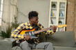 © JustLife - Young joyful african american man playing guitar in living room. Happy man enjoy at home.