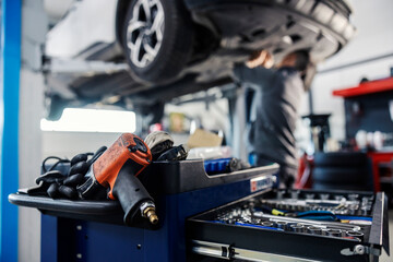 Wall Mural - Selective focus on a drill and drawer with tools on a mechanic toolbox with worker fixing car in blurry background at mechanic's shop.