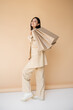 © LIGHTFIELD STUDIOS - full length of cheerful asian woman in stylish pantsuit posing with shopping bags on beige background.