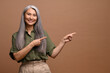 © Vadim Pastuh - Smiling grey haired forty years old asian woman pointing away on blank copy space, showing place for your advertisement isolated over brown background