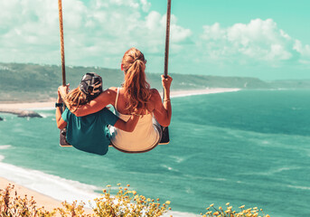 Wall Mural - Mother and son having fun on swing above atlantic ocean- Portugal, Nazare- family love, vacation, relaxing, tourism,tropical beach concept