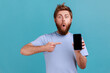 © khosrork - Portrait of astonished shocked bearded man pointing finger at smartphone with black blank screen, copy space for advertisement or promotional text. Indoor studio shot isolated on blue background.