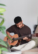 © ADDICTIVE STOCK - Cheerful ethnic man playing guitar with laptop on sofa