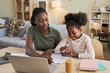 © Seventyfour - African American mother helping her daughter with homework while they sitting at table with laptop in the living room