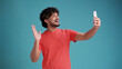 © Виталий Лысак - Young handsome smiling Indian man making video call on the smartphone, waving, chatting via cell phone isolated on blue studio background