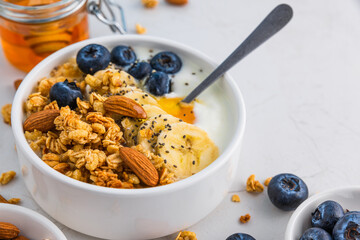 Wall Mural - Bowl of oat granola with yogurt, banana, blueberries, chia seeds, honey and nuts on white background