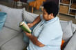© Seventyfour - Side view of senior black woman in t-shirt relaxing on comfortable couch with pillows with book of curious stories or novel in living room