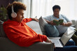 © Seventyfour - Side view of teenage boy in headphones and red hoodie watching online movie or video on laptop screen against woman reading book