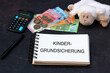 Kindergrundsicherung: Teddybär mit Euro Banknoten, Taschenrechner und dem Text Kindergrundsicherung auf einem Notizblock.	