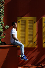 Wall Mural - Handsome young latin man in plain white t-shirt sitting near the rustic house with red walls and yellow window shutters. Close up, copy space, background.