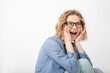 © Татьяна Волкова - Portrait of young surprised woman with open mouth, wearing glasses, denim shirt, sitting, holding cheeks with hands.