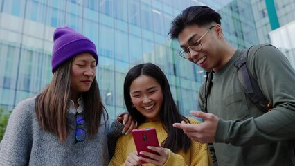 Young adult asian friends having fun looking at mobile phone screen outdoors. Chinese teenage people laughing together watching funny social media content on smart phone network app. 
