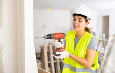 Woman builder in a protective helmet and a yellow vest drills a hole with a screwdriver