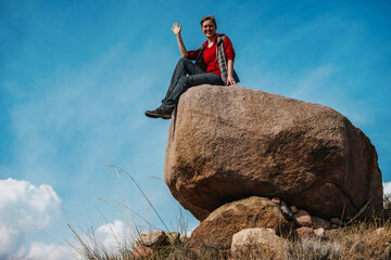 Canvas Print - Young woman tourist sits on a big boulder and waves her hand