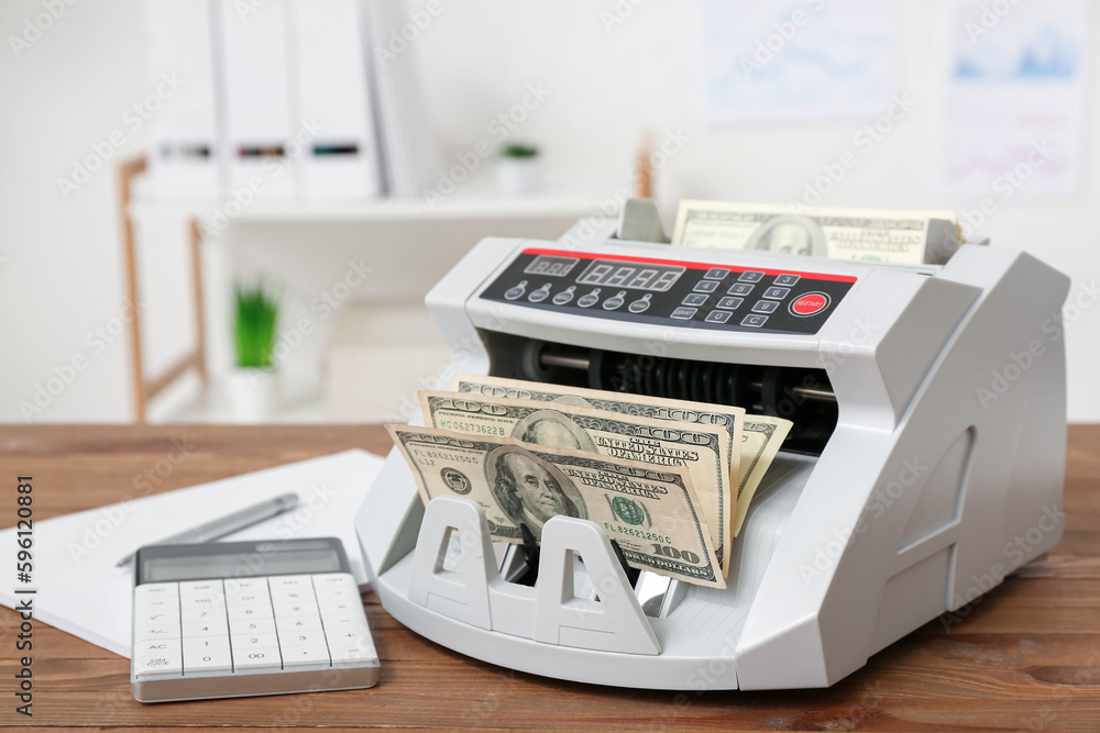 Modern cash counting machine with dollar banknotes and calculator on table in office