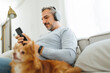 Smiling Bearded mature adult man sitting on the couch wearing headphones listening to music playing in smartphone with his dog. Lifestyle concept, Attractive Middle aged male relax with pet in living 