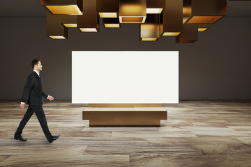 Poster - Side view of businessman walking in modern exhibition room interior with creative ceiling lamps, wooden flooring and empty white mock up banner on concrete wall. Museum and art concept.