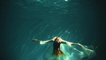Fantasy Woman Drowns Under Dark Blue Water Falls To Bottom Sea. Wet White Silk Long Dress. Red-haired Fairy Girl Goddess River Nymph Mermaid Swims Immersed. Creative Underwater Video Shooting Pool 4k