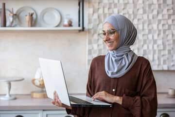 Wall Mural - Joyful and successful business woman in hijab at home using laptop while standing, muslim woman working remotely.