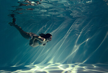 Underwater shoot of beautiful pregnant woman swimming in water through sunbeams.