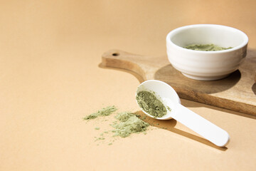 Green powder in a small white ceramic bowl, with a white spoon on a wooden board, on a beige background.  This powder sprinkles the background around.  Super food or marijuana kief concept