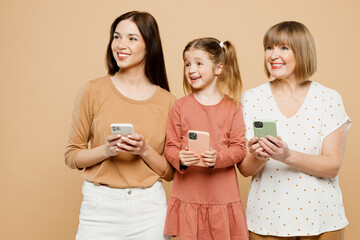 Wall Mural - Happy women wear casual clothes with child kid girl 6-7 years old. Granny mother daughter hold use mobile cell phone look aside on area isolated on plain beige background. Family parent day concept.