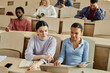 © Seventyfour - Two schoolgirls using laptop together while sitting at desk during lecture with other students in background