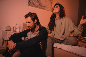 Angry woman sitting near upset boyfriend in bedroom in evening.