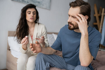 Wall Mural - Bearded man with migraine sitting near blurred girlfriend with pills and water at home.