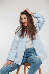 Wall Mural - Funny beautiful fashionable woman with emotions of surprise and joy in trendy denim clothes with a shirt and jeans sits and has fun on a white background in the studio