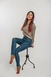 Beautiful young fashionable woman model in fashion casual denim clothes with a vintage sweater and jeans sits and poses on a chair on a white background