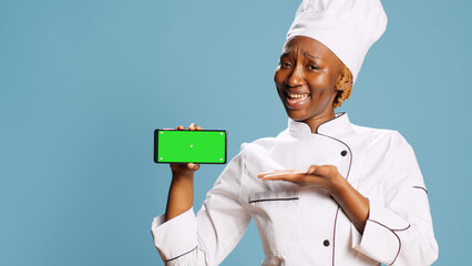 African american chef holding phone with greenscreen, advertising isolated copyspace display in studio. Young woman cook using smartphone with blank chroma key mockup template.