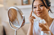 © Drazen - Smiling Asian woman applying face cream while looking herself in mirror.