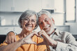 © Daniel - Close up portrait happy sincere middle aged elderly retired family couple making heart gesture with fingers, showing love or demonstrating sincere feelings together indoors, looking at camera..