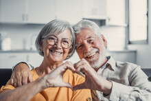 Close Up Portrait Happy Sincere Middle Aged Elderly Retired Family Couple Making Heart Gesture With Fingers, Showing Love Or Demonstrating Sincere Feelings Together Indoors, Looking At Camera..