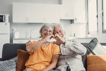 Wall Mural - Close up portrait happy sincere middle aged elderly retired family couple making heart gesture with fingers, showing love or demonstrating sincere feelings together indoors, looking at camera..