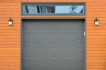 A modern dark brown metal exterior garage door with a small horizontal glass window. The modern door is on a luxury orange contemporary style house with two small light fixtures on either side. 