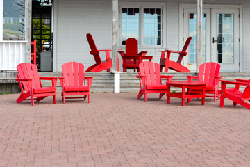 Multiple red painted wooden Adirondack chairs and tables on a restaurant brick patio. There's a white colored building with patio doors, a veranda, and wooden steps.