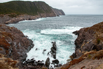 A rugged ocean coastline with high rocky sea stacks, cliffs, vibrant green grass, blue sky, and clouds. The smooth water is a blue color. There's a hiking trail along the edge of the cliff and coast. 