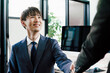 オフィスのパソコンモニターの前で握手をするスーツ姿の20代の若いビジネスマンの男性