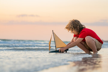 Kid boy playing with toy boat in sea water. Happy holiday by the sea. Kid dreaming about sailing. Happy kid playing with toy sailing boat. Summer vacation. Cute little kid hold toy sailboat.