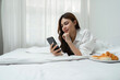 © Natee Meepian - Young woman relaxing and eat bread using telephone on checking social apps and working the bedroom