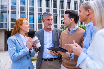 Cheerful group of coworkers laughing and looking at a tablet outdoors in a corporate office area, businesswoman and businessman and executives in commercial area