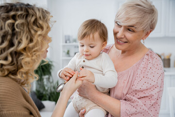 smiling mature woman looking at adult daughter giving spoon to little girl in kitchen.